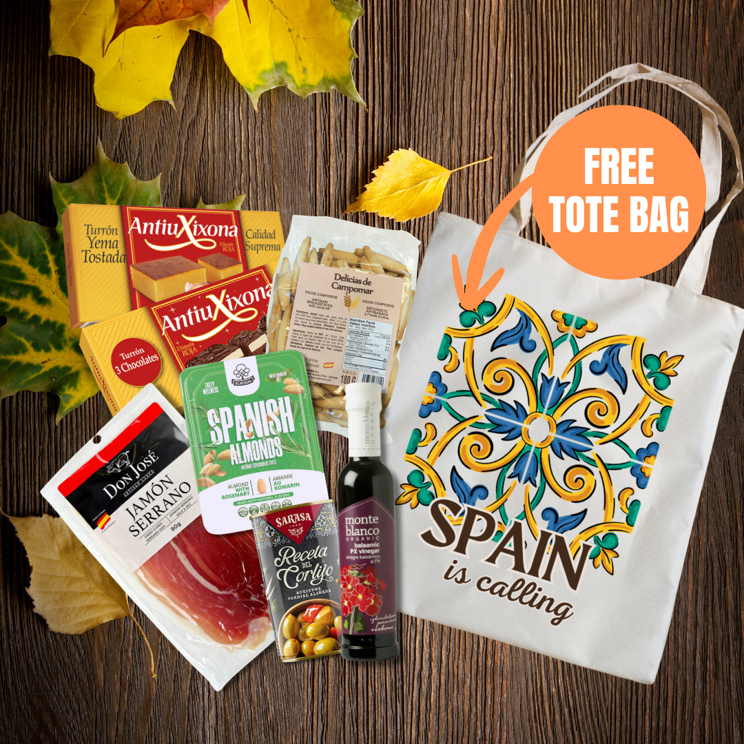 A fall-themed gift basket or subscription box featuring various Spanish snacks and treats, including AntioXona jamón, Sabry Almonds, and a bottle of red wine, arranged with autumn leaves on a dark wood background next to a white tote bag with a colorful tile pattern and the text 'SPAIN is calling'. An orange circle overlay says 'FREE TOTE BAG'.