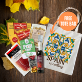 A fall-themed gift basket or subscription box featuring various Spanish snacks and treats, including AntioXona jamón, Sabry Almonds, and a bottle of red wine, arranged with autumn leaves on a dark wood background next to a white tote bag with a colorful tile pattern and the text 'SPAIN is calling'. An orange circle overlay says 'FREE TOTE BAG'.