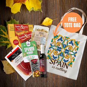 A fall-themed gift basket or subscription box featuring various Spanish snacks and treats, including AntioXona jamón, Sabry Almonds, and a bottle of red wine, arranged with autumn leaves on a dark wood background next to a white tote bag with a colorful tile pattern and the text 'SPAIN is calling'. An orange circle overlay says 'FREE TOTE BAG'.