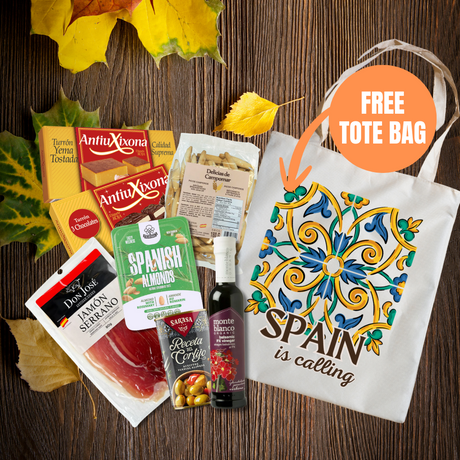 A fall-themed gift basket or subscription box featuring various Spanish snacks and treats, including AntioXona jamón, Sabry Almonds, and a bottle of red wine, arranged with autumn leaves on a dark wood background next to a white tote bag with a colorful tile pattern and the text 'SPAIN is calling'. An orange circle overlay says 'FREE TOTE BAG'.
