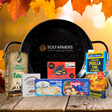 A fall-themed gourmet food gift set arranged on a rustic wooden table with autumn leaves in the background. The set includes a large black speckled pan from Solfarmers, a bag of Sol de la Rama chocolate chips, a box of Colita Blanca Paella Fana, a package of Alamar tortillas, a packet of Saladero Tostadas, and a box of Bogar ground beef pasta.