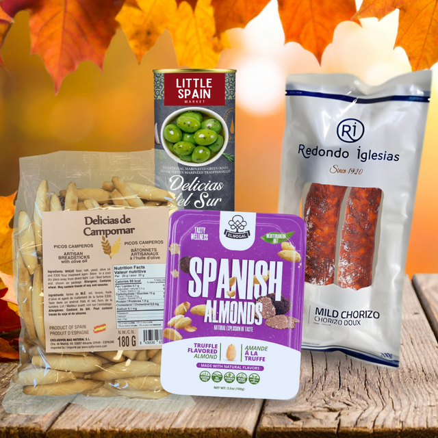 A group of packaged Spanish food products arranged on a rustic wooden surface with autumn leaves in the background. The products include a bag of Campomar pasta, a box of Little Spain green olives, a package of Redondo Iglesias mild chorizo, and a package of Almondaria Spanish Almonds.