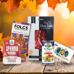 A curated assortment of gourmet food products arranged on a rustic wooden surface with autumn maple leaves and warm bokeh lights in the background. Visible products include a box of Folcs Friendly Chocolate, a package of Don José Ibérico Cebolla Jamón, a Spanish Almonds chocolate-covered almond box, an El Faro Aceitunas can, and a packaged snack item.