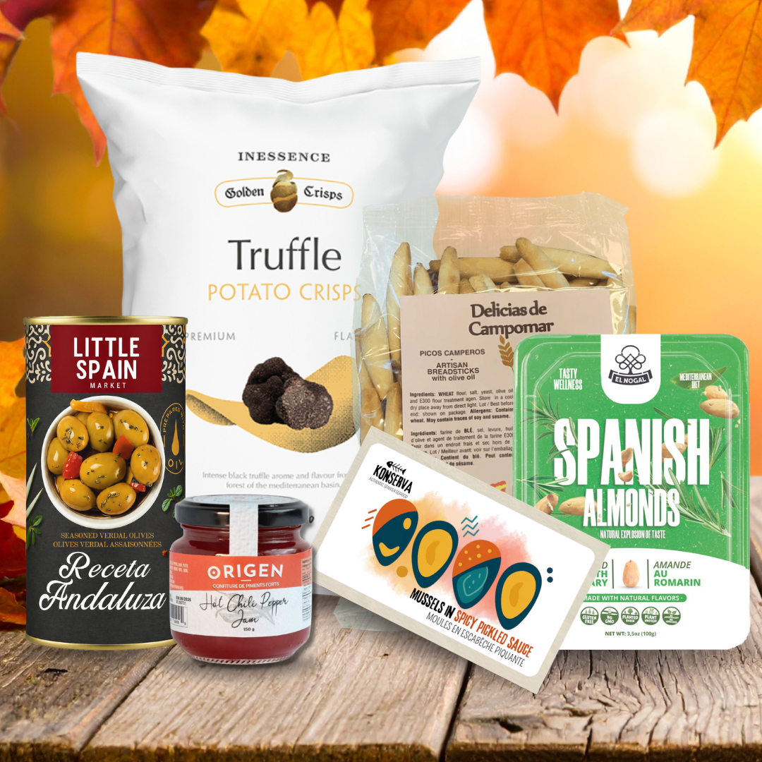 A curated Spanish gourmet gift set arranged on a rustic wooden table with autumn leaves in the background. The set includes a large bag of Inesence Truffle Potato Crisp, a can of Little Spain Olives, a jar of Origen Jamón Ibérico, a package of Delicia de Campomar chips, a pack of Neta Spanish Almonds, and a box of Marigold Gourmet Truffle Cheese.