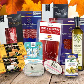 A fall-themed gourmet gift basket featuring an assortment of Spanish snacks and cured meats arranged on a rustic wooden table with autumn leaves in the background. The products include Don José jamon serrano and redondo packages, Anta Brasa crackers, Gourmet Spanish Almonds, Redondo Iglesias jamon, a bottle of olive oil, and a can of El Faro Anchoa.