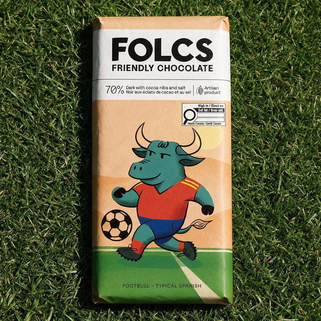 A bar of FOLCS Friendly Chocolate in its packaging, placed on a green grass background. The wrapper features a cartoon illustration of a green bull soccer player wearing a red and blue jersey.