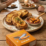 A group of people gathered around a rustic wooden dining table, holding wine glasses and enjoying a meal. In the foreground, a large platter holds toasted bread slices topped with melted cheese and herbs, alongside a small bowl of stuffed olives.