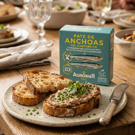 A box of Agromar Paté de Anchoas (cured anchovies) sits on a rustic wooden dining table, styled with slices of toasted bread topped with anchovy spread and fresh chives. In the background, a man and a woman are seated at the table, blurred, creating a warm, family-friendly dining atmosphere.
