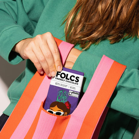 A person wearing a green sweatshirt holds a pink and orange striped tote bag with a pocket, showing a Folcs chocolate bar tucked inside.